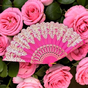 May include: A bright pink folding hand fan featuring intricate gold-colored lace patterns along the upper edge and a decorative filigree design on the lower ribs, displayed against a background of multiple blooming pink rose flowers.