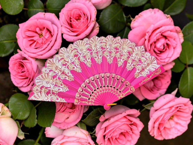 May include: A bright pink folding hand fan featuring intricate gold-colored lace patterns along the upper edge and a decorative filigree design on the lower ribs, displayed against a background of multiple blooming pink rose flowers.