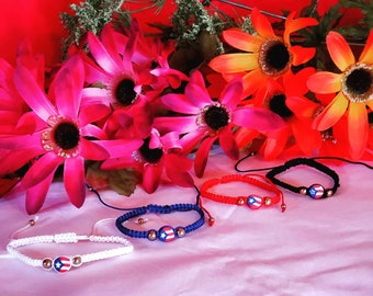 May include: Five handmade bracelets with the Puerto Rican flag design. The bracelets are in white, blue, red, and black, with gold-colored beads. The background features bright pink and orange flowers against a red backdrop.