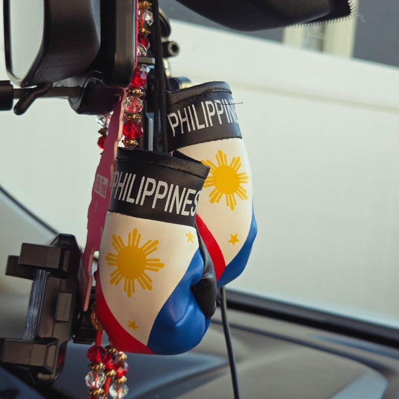 Puede incluir: Peque&ntilde;os guantes de boxeo con la palabra "PHILIPPINES" impresa. Los guantes son blancos, azules y rojos, con un dise&ntilde;o de sol amarillo y estrellas. Cuelgan del espejo retrovisor de un coche.
