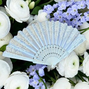May include: A light blue folding hand fan featuring an intricate white lace pattern across the fabric surface and a matching light blue plastic frame with decorative cutouts at the base, displayed in an open, semi-circular position.