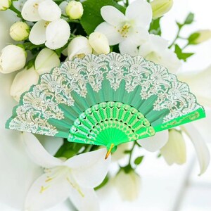 May include: A bright green folding hand fan featuring an intricate silver lace overlay pattern on the upper portion and a solid green plastic frame with a decorative cutout design on the lower section, held together by a small gold-toned metal rivet.