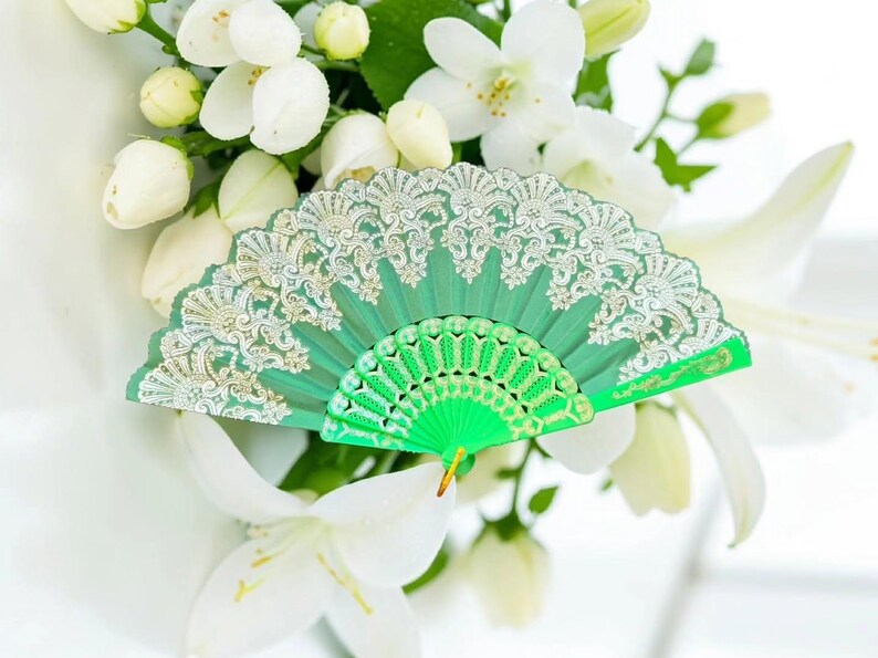 May include: A bright green folding hand fan featuring an intricate silver lace overlay pattern on the upper portion and a solid green plastic frame with a decorative cutout design on the lower section, held together by a small gold-toned metal rivet.