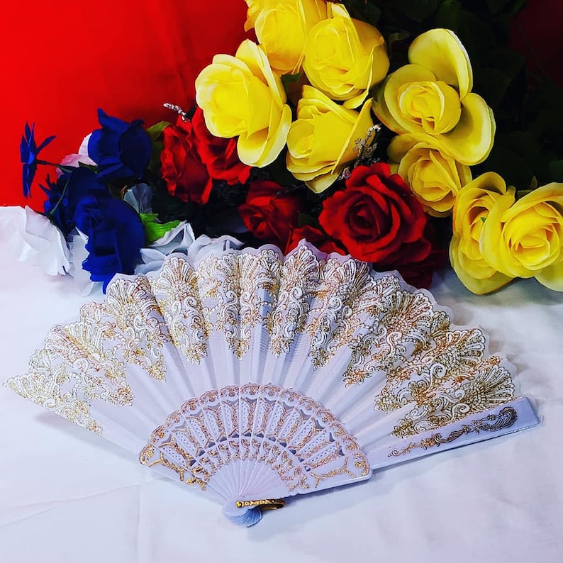 May include: A white folding hand fan featuring intricate gold-colored lace detailing along the top edge and a decorative gold pattern on the lower ribs, positioned in front of a bouquet of yellow, red, and blue artificial roses.