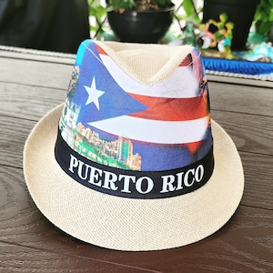 May include: A straw hat with a black band that says "PUERTO RICO" in white letters. The hat is decorated with a colorful image of the Puerto Rican flag and a cityscape.