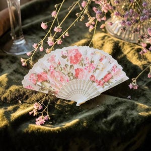 May include: A folding hand fan featuring a white fabric leaf printed with a detailed pattern of pink roses and green leaves. The fan has a white plastic frame with intricate embossed detailing on the ribs and a scalloped edge along the top.