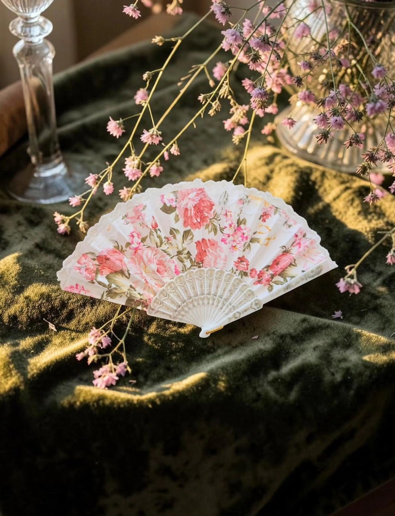 May include: A folding hand fan featuring a white fabric leaf printed with a detailed pattern of pink roses and green leaves. The fan has a white plastic frame with intricate embossed detailing on the ribs and a scalloped edge along the top.
