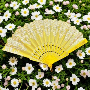 May include: A bright yellow folding hand fan featuring an intricate white lace pattern along the top edge and a solid yellow plastic frame with decorative cutouts at the base, displayed in a fully open, semi-circular position.