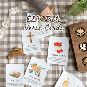 May include: ESV Bible Verse & Cards with watercolor illustrations of a cross, strawberries, eggs, a wheelbarrow, honey, and a plant. The cards are arranged on a checkered surface with wooden animal figurines and a wooden tray.