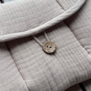 May include: Close-up of a light beige, quilted fabric pouch with a rounded flap. The pouch is secured with a wooden button and a fabric loop. The fabric has a textured appearance.