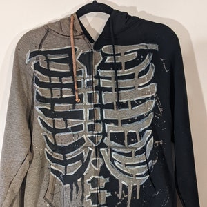 The front view of a zip up hooded sweater. The left half of the sweater has been bleached gray, and the right half is black. A dripping skeleton ribcage outlined in white is painted black on the gray side and bleached gray on the black side.