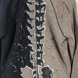 A close up of the back of a hooded zip up sweater. The left half of the sweater is black while the right half has been bleached gray. A skeleton spine has been painted black on the gray side and is gray on the black side.