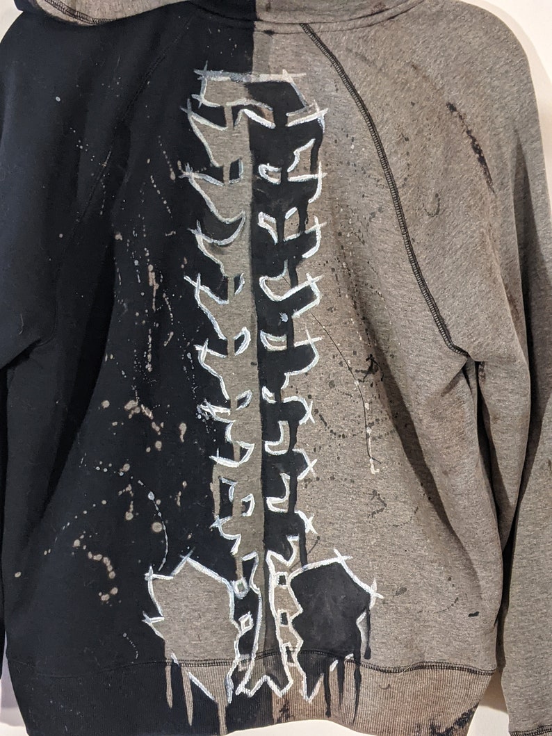 A close up of the back of a hooded zip up sweater. The left half of the sweater is black while the right half has been bleached gray. A skeleton spine has been painted black on the gray side and is gray on the black side.