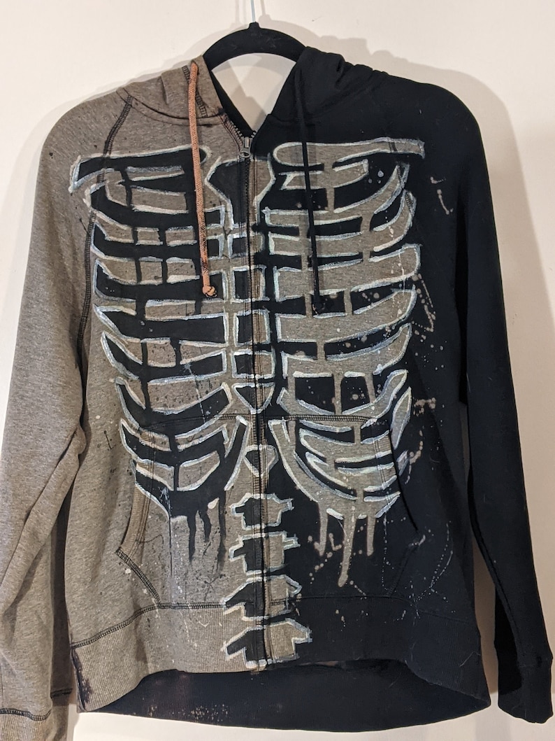 The front view of a zip up hooded sweater. The left half of the sweater has been bleached gray, and the right half is black. A dripping skeleton ribcage outlined in white is painted black on the gray side and bleached gray on the black side.