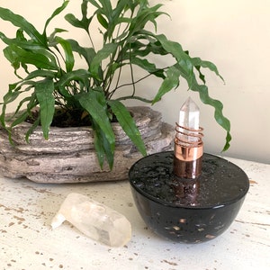 Puede incluir: Una botella de vidrio negro con motas doradas y una tapa de cristal de cuarzo transparente. La botella está sobre una superficie blanca con un pequeño cristal de cuarzo transparente al lado.