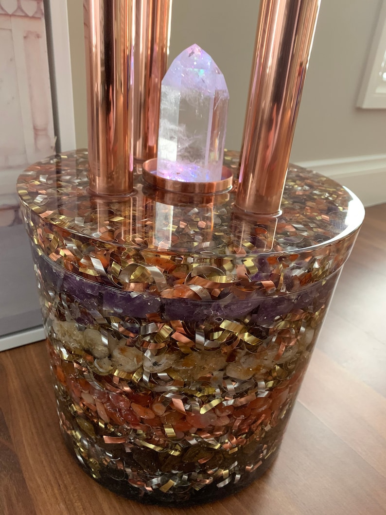 Puede incluir: Un orbe de vidrio transparente lleno de capas de cristales de diferentes colores y tuber&iacute;as de cobre. El orbe contiene varios cristales, incluyendo amatista, citrino y cuarzo rosa. Las tuber&iacute;as de cobre est&aacute;n dispuestas en forma triangular alrededor del orbe.