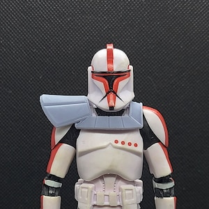 May include: A white and red Star Wars action figure with a blue shoulder pad. The figure is wearing a helmet with a red visor and a white and red chest plate.