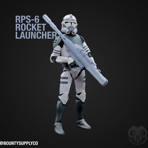 Op de afbeelding: Een wit en grijs action figure van een Star Wars kloon soldaat die een grijze RPS-6 raketwerper vasthoudt. De figuur heeft een witte helm met een zwart vizier en zwarte markeringen op het pantser. De tekst "RPS-6 ROCKET LAUNCHER" staat in de linkerbovenhoek van de afbeelding.