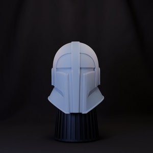 Puede incluir: Un casco blanco con un diseño futurista, exhibido en un soporte negro. El casco tiene una forma lisa y redondeada con una cresta elevada en el centro.