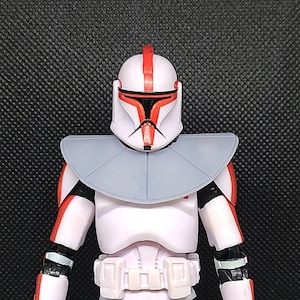 Peut inclure: Une figurine d'action blanche et rouge d'un clone trooper de Star Wars avec un casque gris et un col gris. La figurine porte une armure blanche et rouge avec une ceinture noire.