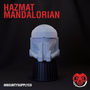 May include: A light blue Hazmat Mandalorian helmet, displayed on a black base. The text "HAZMAT MANDALORIAN" is in red at the top. The @BOUNTYSUPPLYCO logo is at the bottom. The helmet has a detailed design.