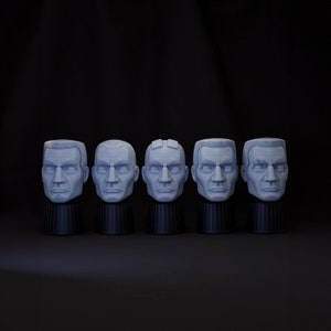 May include: Five gray 3D printed busts of a man's head, each with a slightly different facial expression. The busts are arranged in a row on a black surface.