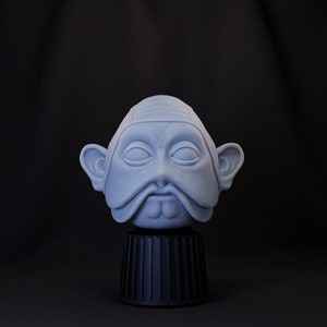 May include: A white ceramic bust of a person with large ears, a mustache, and a goatee. The bust is on a black pedestal.