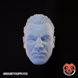 May include: A light blue bust of a man's head with detailed facial features and textured skin. The left side of the face has a scarred appearance. The image includes the text "@BOUNTYSUPPLYCO" at the bottom left and a logo at the bottom right.