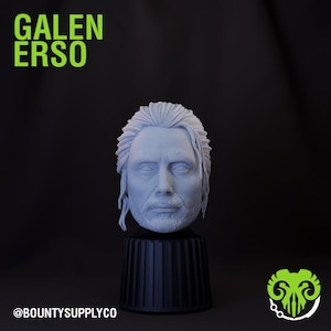 May include: A 3D printed bust of Galen Erso, a character from the Star Wars franchise. The bust is painted in a light grey colour and is displayed on a black pedestal. The text "GALEN ERSO" is displayed in green letters at the top of the image. The text "@BOUNTYSUPPLYCO" is displayed at the bottom of the image.