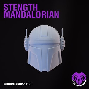 Puede incluir: Un casco mandaloriano azul claro con el texto "STENGTH MANDALORIAN" en morado. El casco tiene estructuras tipo antena en los lados y está sobre un fondo negro. El logotipo @BOUNTYSUPPLYCO está en la parte inferior.