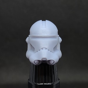 Puede incluir: Un casco de soldado clon blanco de plástico con una visera negra. El casco está sobre una superficie negra.