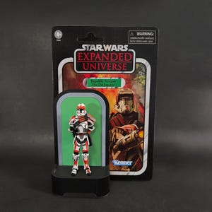 May include: A Star Wars Expanded Universe Republic Trooper action figure, displayed in a custom stand. The figure is white, red, and black, with a helmet. The packaging features the text "Star Wars Expanded Universe" and "Kenner".