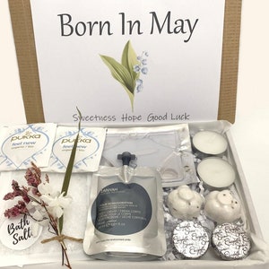 May include: A gift box with the text "Born In May" and a floral illustration. The box contains tea bags, bath salts, a body cream, candles, and chocolates. The box is filled with white tissue paper.