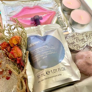 May include: A gift box containing beauty and self-care items. Includes a sachet of shampoo, lip mask, two small candles, two wrapped soaps, and two pink bath bombs. A small bouquet of dried flowers and rose petals are also included.
