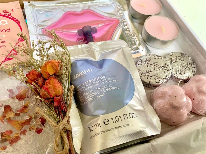 May include: A gift box containing beauty and self-care items. Includes a sachet of shampoo, lip mask, two small candles, two wrapped soaps, and two pink bath bombs. A small bouquet of dried flowers and rose petals are also included.