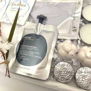 May include: A gift box containing spa products. Includes tea packets, body cream, candles, wrapped soaps, and chocolates. The box is lined with white paper and decorated with dried flowers and greenery.