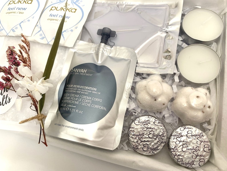 May include: A gift box containing spa products. Includes tea packets, body cream, candles, wrapped soaps, and chocolates. The box is lined with white paper and decorated with dried flowers and greenery.