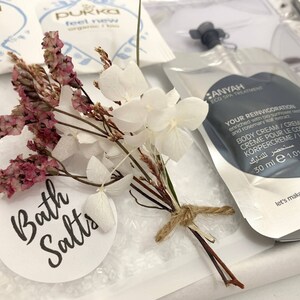 May include: A spa-themed gift set featuring bath salts, dried flowers, and body cream. The set includes a small bouquet of white and pink dried flowers tied with twine. The bath salts are in a clear bag with a label that reads "Bath Salts".