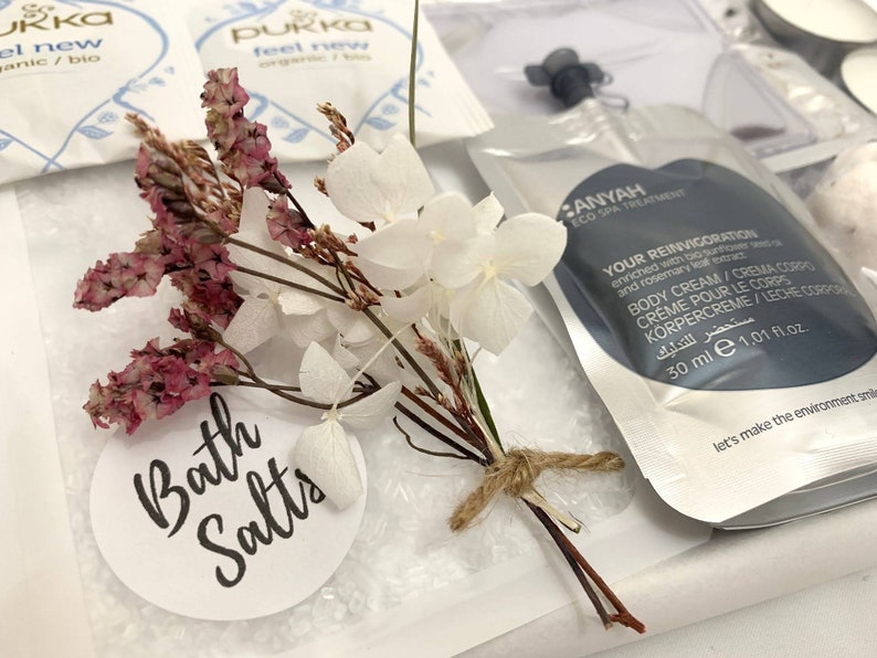 May include: A spa-themed gift set featuring bath salts, dried flowers, and body cream. The set includes a small bouquet of white and pink dried flowers tied with twine. The bath salts are in a clear bag with a label that reads "Bath Salts".