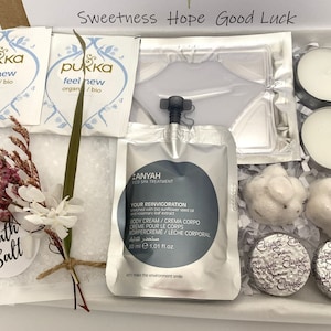 May include: A gift box filled with self-care items. Includes two tea bags, a face mask, two tea light candles, two bath bombs, two wrapped chocolates, bath salts, and a small floral arrangement. The box has the words "Sweetness Hope Good Luck".