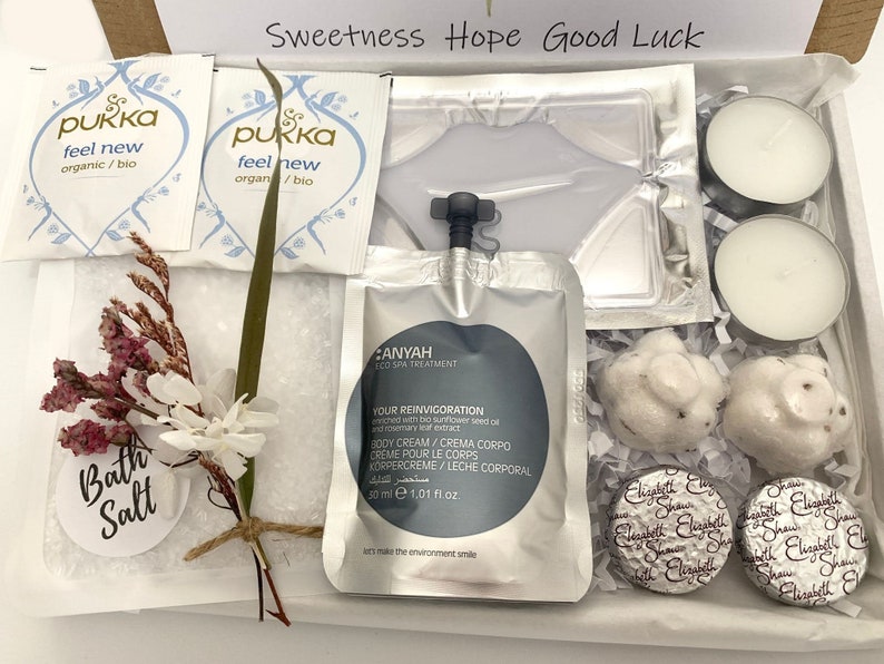 May include: A gift box filled with self-care items. Includes two tea bags, a face mask, two tea light candles, two bath bombs, two wrapped chocolates, bath salts, and a small floral arrangement. The box has the words "Sweetness Hope Good Luck".