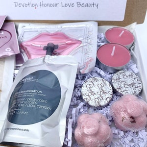 Pu&ograve; includere: Una confezione regalo con prodotti di bellezza, tra cui candele rosa, bombe da bagno e prodotti per la cura della pelle. La scatola contiene una maschera per le labbra, crema corpo e cioccolatini confezionati. Il testo "Devotion Honour Love Beauty" &egrave; visibile.