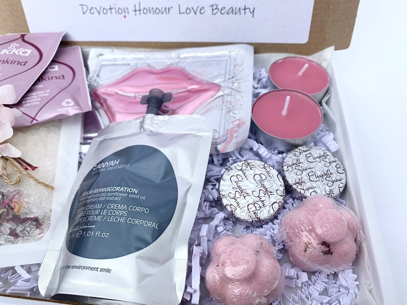 Pu&ograve; includere: Una confezione regalo con prodotti di bellezza, tra cui candele rosa, bombe da bagno e prodotti per la cura della pelle. La scatola contiene una maschera per le labbra, crema corpo e cioccolatini confezionati. Il testo "Devotion Honour Love Beauty" &egrave; visibile.