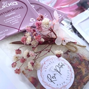 Pu&ograve; includere: Primo piano di un set regalo con una bustina di sali da bagno, una bustina di t&egrave; e fiori secchi. La bustina di t&egrave; &egrave; etichettata "Pukka womankind organic". I sali da bagno sono in un sacchetto trasparente con un'etichetta "Natural Rose Petal Bath Salts".