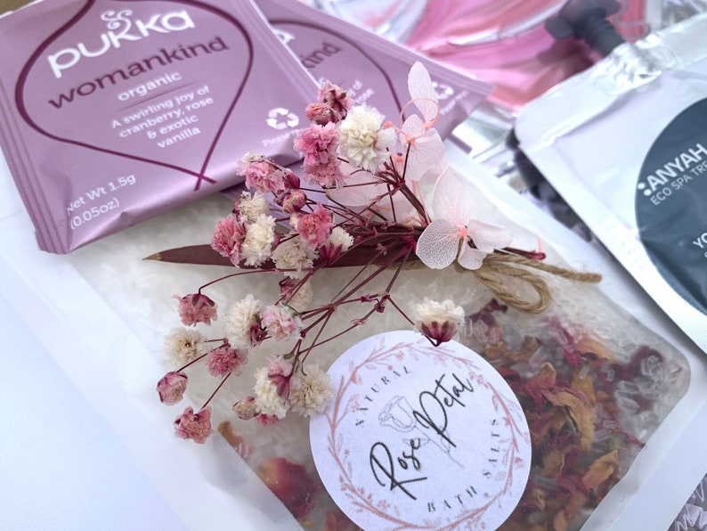 Pu&ograve; includere: Primo piano di un set regalo con una bustina di sali da bagno, una bustina di t&egrave; e fiori secchi. La bustina di t&egrave; &egrave; etichettata "Pukka womankind organic". I sali da bagno sono in un sacchetto trasparente con un'etichetta "Natural Rose Petal Bath Salts".