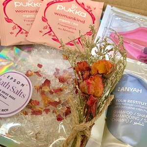 May include: A gift box containing bath products. Includes Epsom bath salts with dried rose petals, two packets of Pukka womankind tea, a bouquet of dried flowers, and a shampoo sachet. The box also contains a pink lip-shaped item.