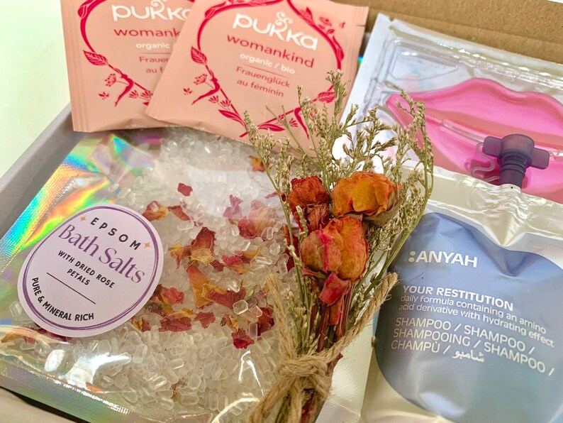 May include: A gift box containing bath products. Includes Epsom bath salts with dried rose petals, two packets of Pukka womankind tea, a bouquet of dried flowers, and a shampoo sachet. The box also contains a pink lip-shaped item.