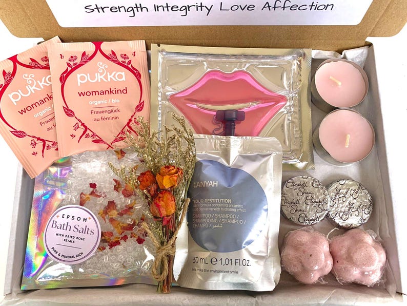 May include: A gift box filled with self-care items. Includes Pukka tea packets, a lip-shaped face mask, bath salts with rose petals, a small bouquet of dried flowers, shampoo, two candles, two bath sponges, and two wrapped soaps.