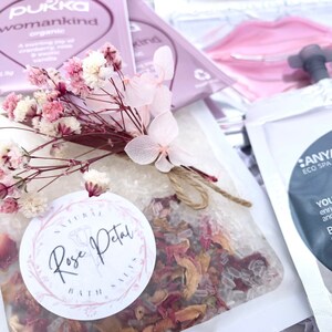 Pu&ograve; includere: Una collezione di prodotti per la cura di s&eacute;, tra cui una bustina di sali da bagno ai petali di rosa, una bustina di t&egrave; e una bustina di crema corpo. L'immagine presenta un mazzo di fiori rosa e bianchi secchi legati con spago.