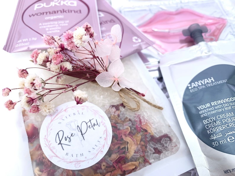 Pu&ograve; includere: Una collezione di prodotti per la cura di s&eacute;, tra cui una bustina di sali da bagno ai petali di rosa, una bustina di t&egrave; e una bustina di crema corpo. L'immagine presenta un mazzo di fiori rosa e bianchi secchi legati con spago.